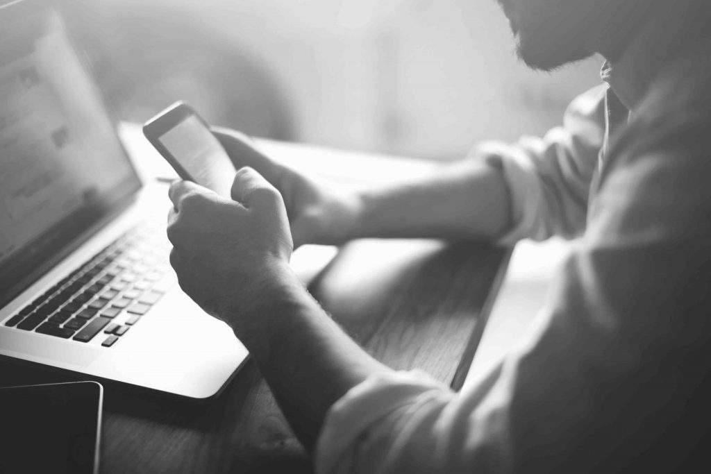 A man reading content on his smart phone with a computer next to him