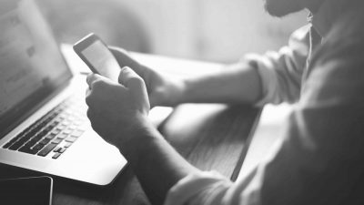 A man reading content on his smart phone with a computer next to him